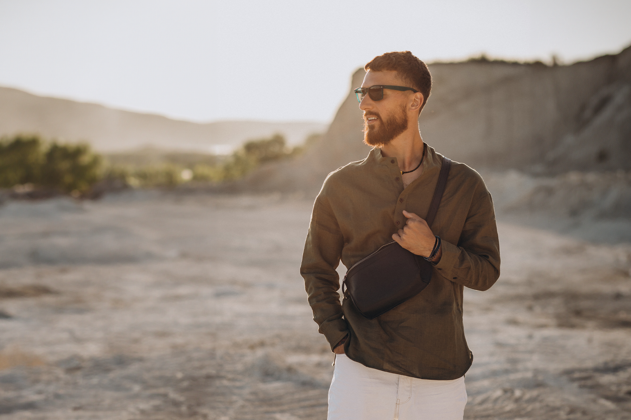 A man wearing sunglasses and a fanny pack stands in a vast desert landscape under a clear blue sky.